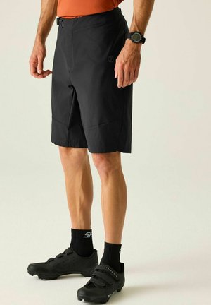 Mann trägt schwarze Fahrradschuhe, schwarze Shorts, schwarze Socken und ein oranges Hemd und steht vor einem einfarbigen Hintergrund.