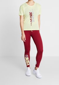 Maglietta in rete giallo chiaro con grafica laterale, abbinata a leggings bordeaux aderenti con un grande logo e accenti bianchi. Scarpe bianche.