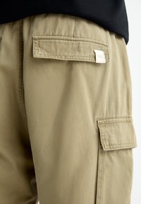 Pantalones de cargo beige con dos bolsillos laterales, tejido texturizado y una pequeña etiqueta. Presentan un ajuste relajado y cintura elástica.
