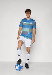 Blauwe en groene gestreepte sporttrui met een ronde hals, gecombineerd met witte shorts en zwarte voetbalschoenen, tegen een effen grijze achtergrond.