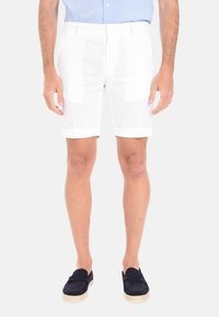 Shorts bianchi in lino con risvolti, caratterizzati da una texture leggera. Indossati con scarpe slip-on blu. Il modello è in piedi su uno sfondo neutro.