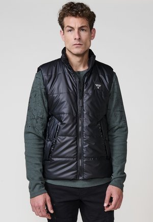 Chaleco puffy negro con un cuello alto, bolsillos con cremallera a la derecha e izquierda y un logo. Lleva puesto sobre una camiseta de manga larga verde oscuro con un estampado.