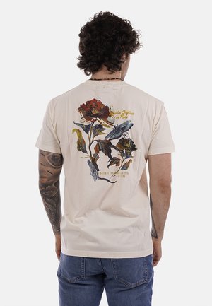 Uomo con capelli ricci e tatuaggi indossa una maglietta beige con una grande stampa floreale e di insetti sul retro, abbinata a jeans blu.