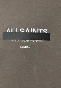 Oliwkowa zieleń tkaniny z wypukłym białym napisem logo "ALL SAINTS" i "LONDON", z czarnym prostokątnym nakładką częściowo zasłaniającą tekst.