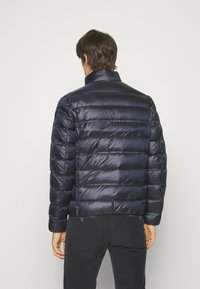 La giacca a vento blu navy presenta un motivo a righe orizzontali, colletto alto e una finitura lucida. Il materiale sembra leggero e isolante.