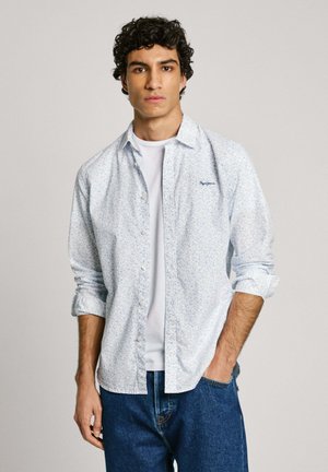 Chemise bleu clair à manches longues avec un petit motif floral, portée avec un t-shirt blanc en dessous et un jean en denim bleu. Manches retroussées.