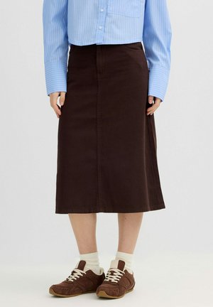 Persona con falda midi marrón, zapatillas marrones con cordones blancos, calcetines blancos y una camisa de manga larga azul con rayas verticales.