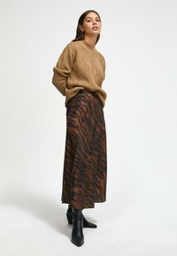 Pull en tricot marron avec une texture détaillée associé à une jupe longue ornée d'un motif rayé tigre en marron foncé et noir. Bottes à talons noires.
