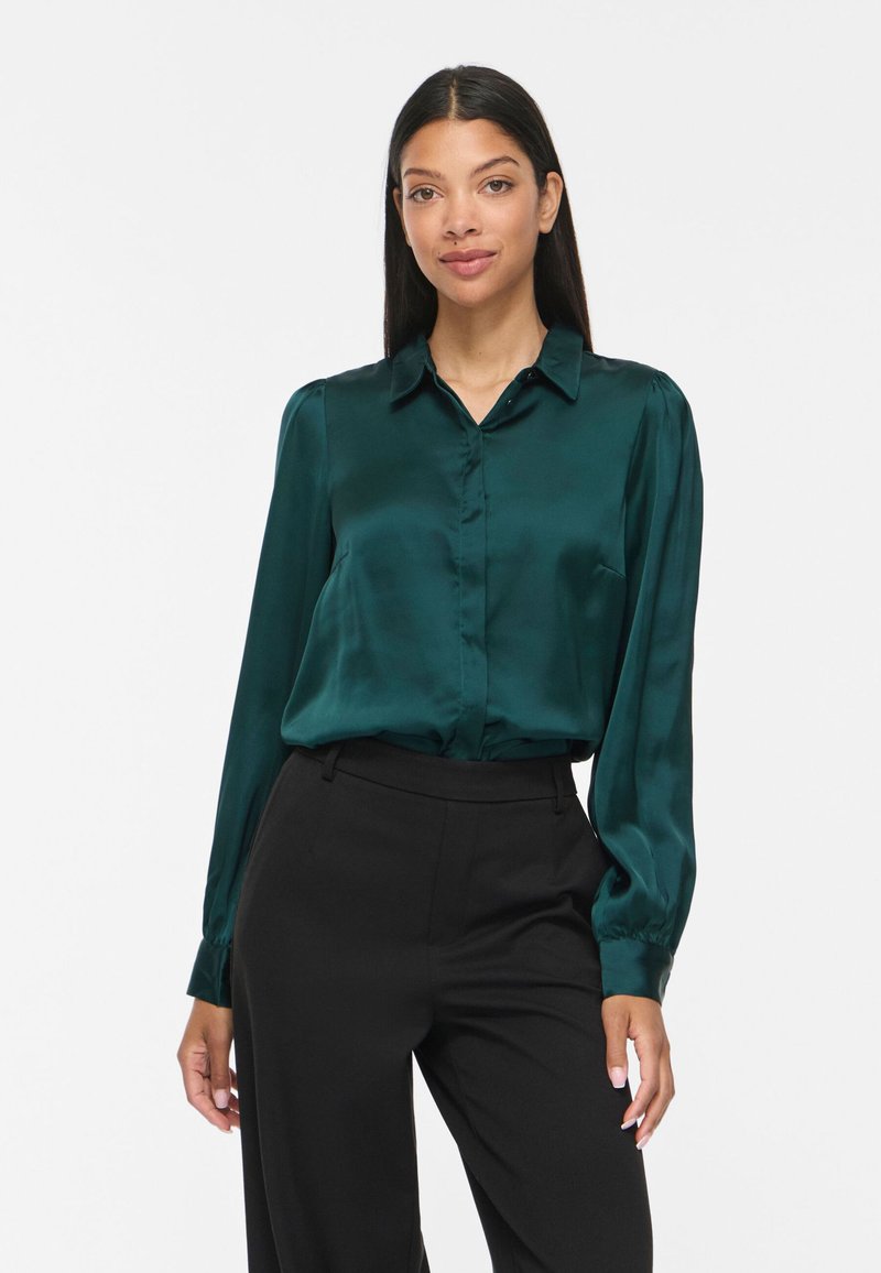 Femme portant un chemisier en satin vert foncé à manches longues, rentré dans un pantalon noir taille haute, debout devant un fond clair uni.