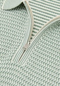 Nahaufnahme eines grünen und weißen texturierten Stoffs mit einem teilweise geöffneten silbernen Reißverschluss.