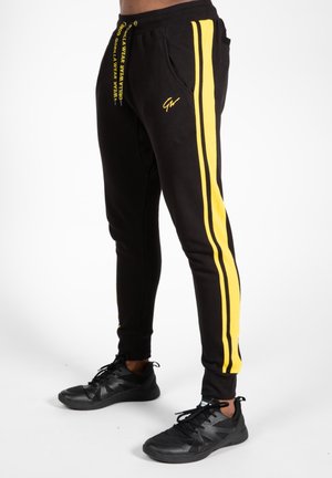 Black joggers with yellow side stripes, tapered fit, drawstring waist, and pockets. Paired with black athletic shoes.