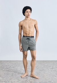 Szürke fürdőnadrág elasztikus derékrésszel, fekete húzózsinórokkal és logóval. A modell mezítláb áll, előre nézve, világos háttér előtt.