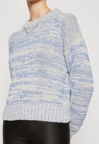 Gebreide sweater in lichtblauw en crème met een textuurpatroon, ronde halslijn en geribde zoom. Draagt over zwarte strakke broek.
