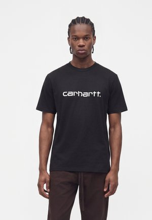 Νέος άνδρας με πλεξούδες, που φοράει μαύρο T-shirt Carhartt και σκούρο παντελόνι, στέκεται μπροστά σε έναν απλό λευκό φόντο.