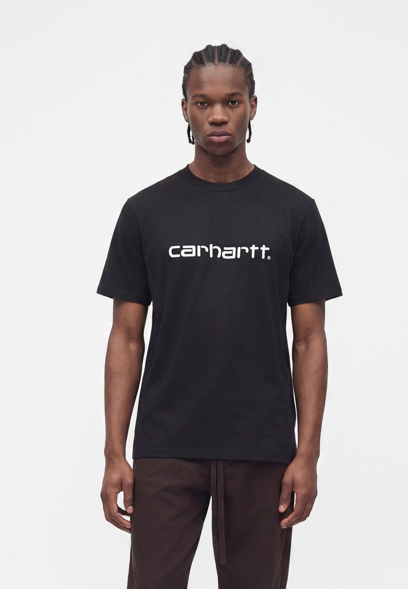Νέος άνδρας με πλεξούδες, που φοράει μαύρο T-shirt Carhartt και σκούρο παντελόνι, στέκεται μπροστά σε έναν απλό λευκό φόντο.