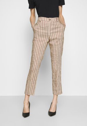 Persoon draagt beige cropped broek met verticale rode en zwarte strepen, zwarte top en zwarte puntige hakken, staand met handen in de zakken.