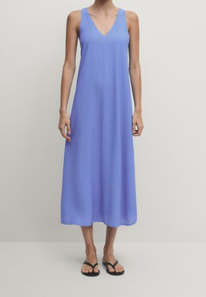 Robe midi lavande sans manches avec col en V portée avec des tongs noires sur une personne debout devant un fond uni.