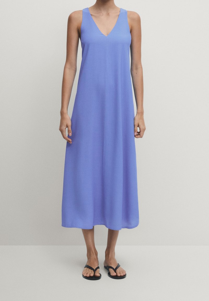Robe midi lavande sans manches avec col en V portée avec des tongs noires sur une personne debout devant un fond uni.