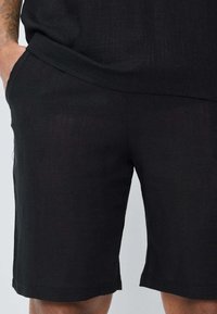 Shorts en lin noir avec une coupe droite, des poches latérales et une texture douce. Le tissu présente un motif de tissage subtil.