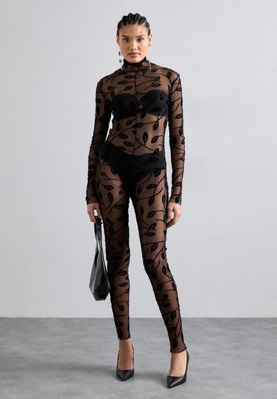 Black sheer mesh bodysuit with leaf patterns, long sleeves, mock neck, and matching short undergarment. Model holding a black tote bag.