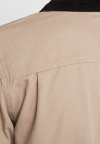 Beige-kankainen kangas, jossa näkyvät ompeleet ja pleat-yksityiskohdat mustan kauluksen lähellä vaatekappaleessa.