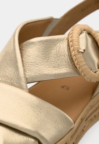 Gros plan d'une sandale en cuir beige avec des larges brides et une boucle tissée ronde sur une semelle plateforme en bois.