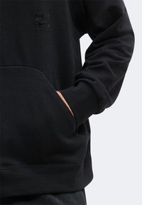 Sweat-shirt noir à manches longues avec poche avant et logo Nike embossé discret sur la poitrine, avec la main partiellement dans la poche.