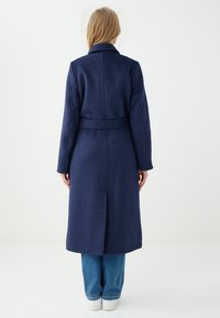 Marineblauwe wollen jas met lange mouwen, een ceintuurlijn en een rugventilatie. Heeft een klassieke kraag en een gladde textuur.