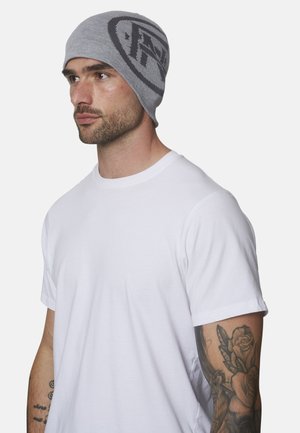 Hombre con barba corta que lleva un gorro gris con patrón negro y una camiseta blanca, mostrando un tatuaje floral en el antebrazo derecho.