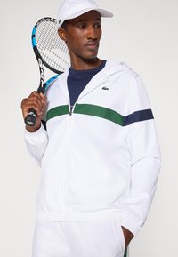 Casaco branco com fecho zip, listras verdes e azuis marinho, punhos e cós elásticos. Modelo segura uma raquete de ténis, veste um boné branco e camisola azul marinho.