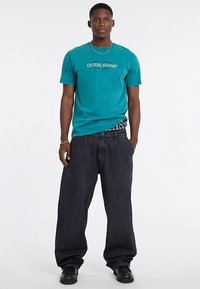 Turquoise katoenen T-shirt met "GUESS JEANS" logo, gecombineerd met loszittende zwarte denim jeans. Model draagt zwarte schoenen.
