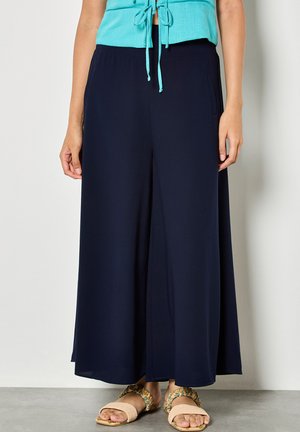 Femme portant un pantalon large marine, un haut turquoise sans manches avec des liens à l'avant, et des sandales beiges à brides florales, debout contre un mur blanc.