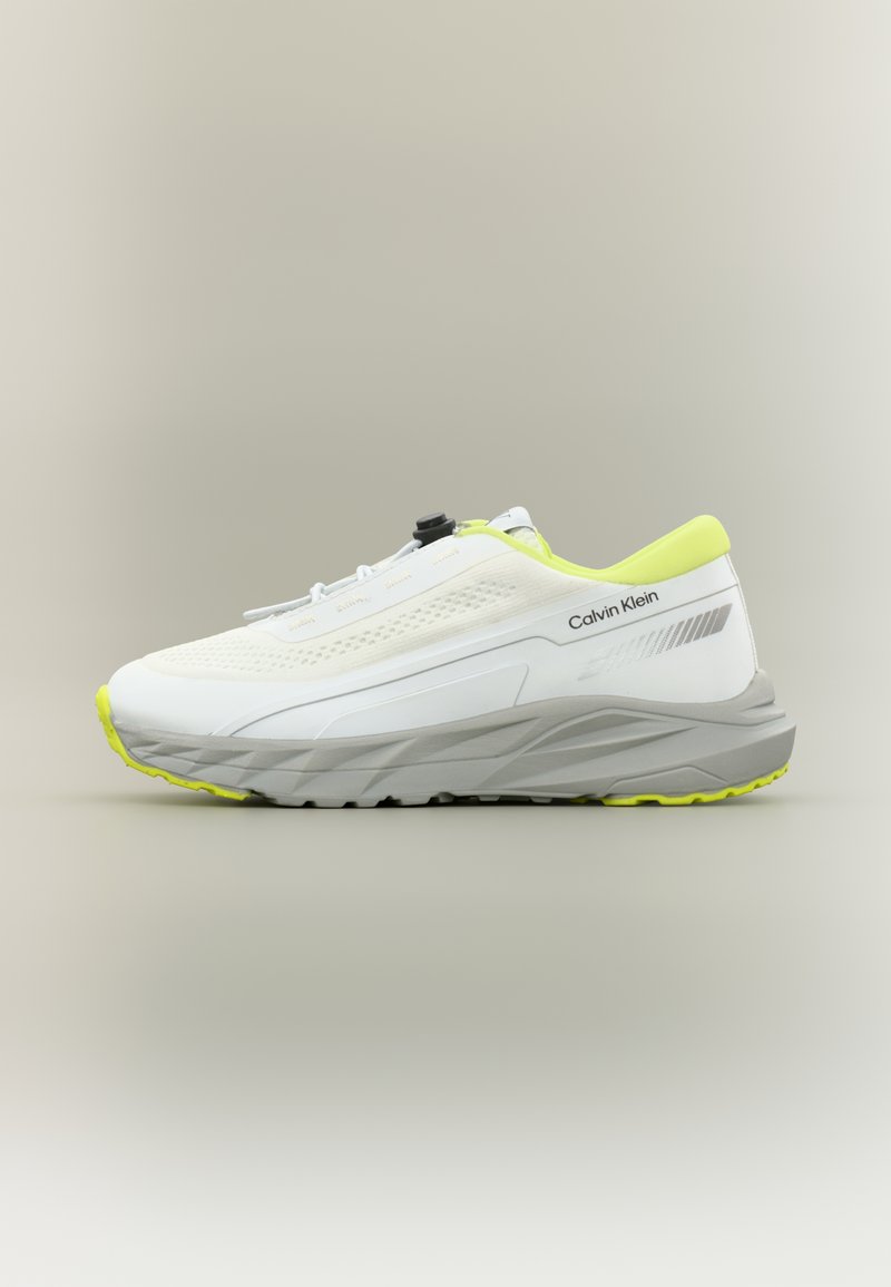 Λευκό αθλητικό παπούτσι με επάνω μέρος από δίχτυ, στρογγυλεμένη μύτη και ασφαλές σύστημα κλειδώματος κορδονιών. Διαθέτει έντονες κίτρινες λεπτομέρειες και γκρι σόλα με πέλμα για καλύτερη πρόσφυση.