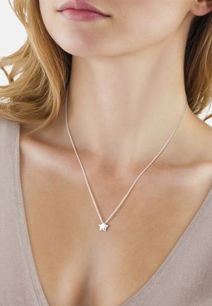 Nærbillede af en kvinde iført en sart sølvstjerneformet vedhæng halskæde på en fin kæde, mod en lysebeige baggrund med top.