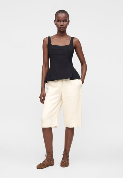 Haut péplum noir sans manches associé à un short crème longueur genou et des ballerines marron à lacets portés par une femme debout sur un fond uni.