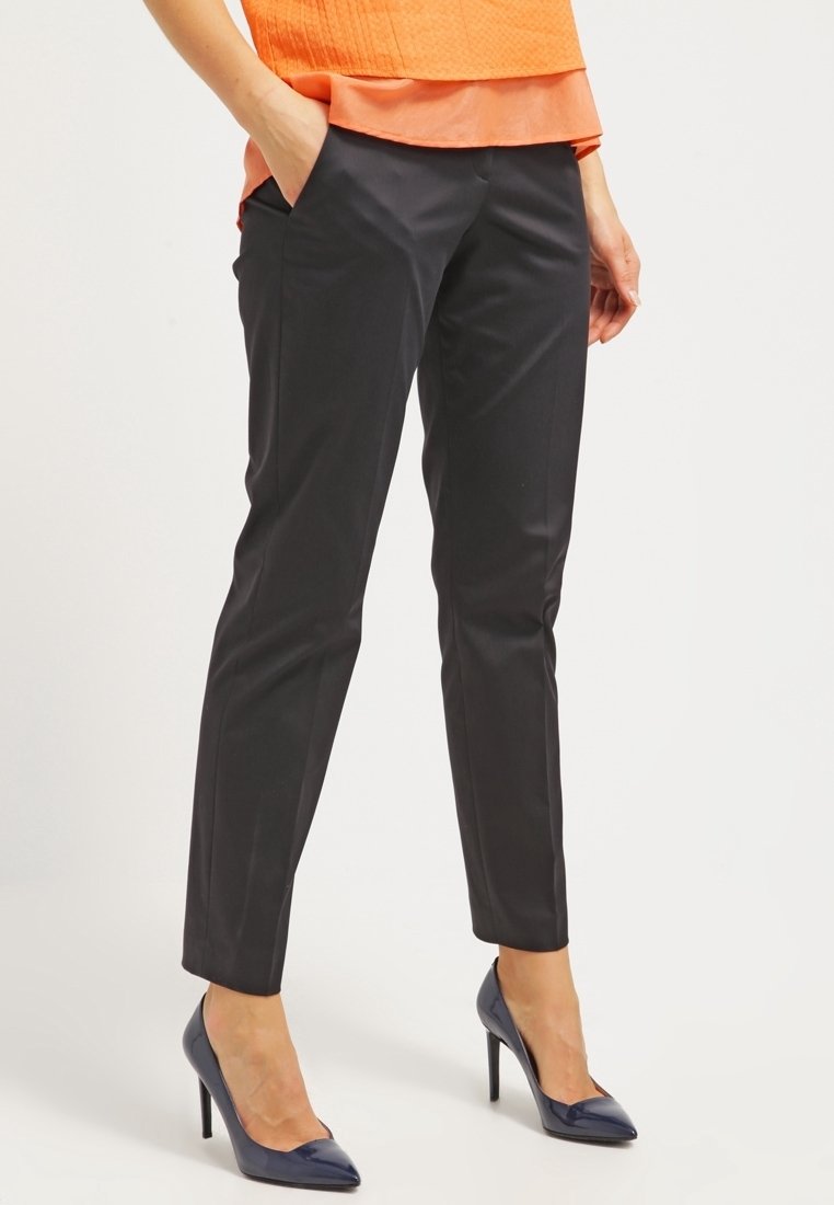 Femme portant un pantalon noir ajusté et des escarpins bleu marine, debout avec une main dans la poche devant un fond uni.