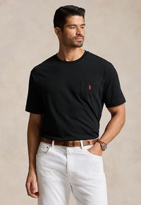 Man draagt een zwart shirt met korte mouwen met rood logo op zak, witte broek, bruine riem en horloge, staand met één hand in de zak.