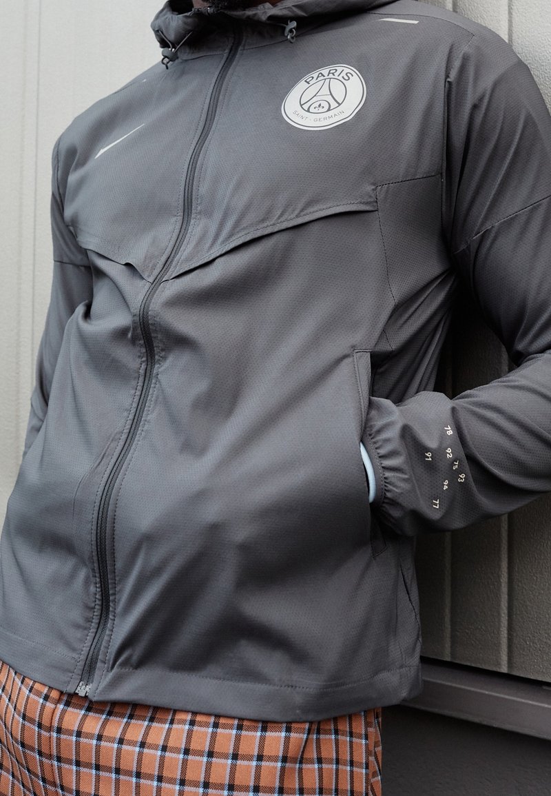 Personne portant une veste Paris Saint-Germain gris foncé avec une fermeture éclair à l’avant et un pantalon à motifs, appuyée contre un mur avec une main dans la poche.