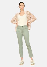 Lichte, gemusterde bomberjack over een strakke witte tanktop; gecombineerd met zachtgroene broek tot de enkels en beige sneakers.