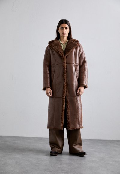 Person standing against plain wall wearing a long brown leather coat with shearling trim, wide-legged pants, and dark shoes.