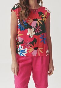 Oberteil mit Blumenmuster in Rot, mit kurzen Ärmeln und rundem Ausschnitt; lebendige Farben wie Pink, Blau und Weiß auf grünem Hintergrund.
