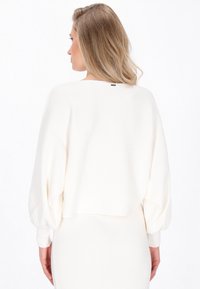 Pull en tricot blanc à côtes avec des manches ballons, design court et un subtil accent noir au niveau de l'encolure arrière. Texture douce.