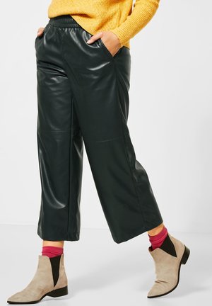 Pantaloni a gamba larga in ecopelle nera con tasche laterali, caratterizzati da una texture liscia e una vita elastica, abbinati a stivali alla caviglia beige e calzini rosa.