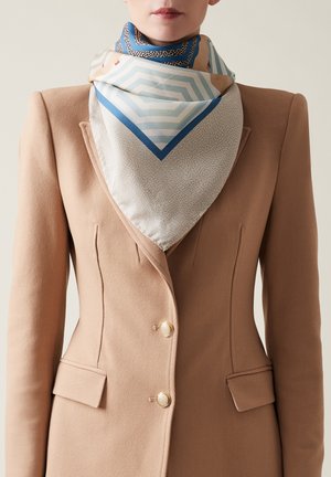 Foulard quadrato in seta con motivi geometrici in blu, crema e beige. I bordi presentano dettagli intricati. Indossato con un blazer beige.