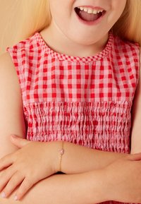 Rode en roze geruite jurk met een gesmockt lijfje; de pols van het kind versierd met een gouden kettingarmband met een roze hartje.