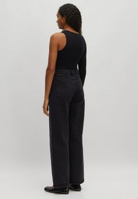 Zwart geribbeld body met één lange mouw, gecombineerd met hoogwaist donker jeans. Model draagt zwarte flats en staat voor een neutrale achtergrond.