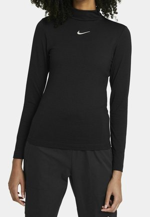 Femme portant un t-shirt noir à manches longues et col roulé Nike avec un pantalon noir sur un fond gris clair.