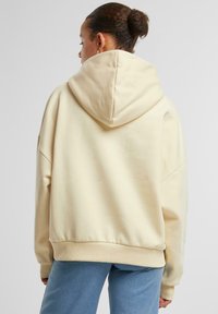 Felpa oversized color crema con un ampio cappuccio arrotondato e polsini a costine. Tessuto morbido con una vestibilità rilassata. Abbinata a pantaloni azzurri.