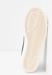 Semelle de chaussure avec un motif en chevron texturé, dotée d'une base en caoutchouc blanc et d'un bord supérieur sombre. Positionnée verticalement sur un fond clair.