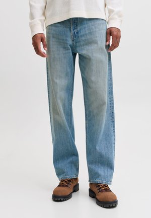 Homme portant un jean large bleu clair, des chaussures de randonnée marron avec des semelles noires et un haut blanc à manches longues, debout sur un fond uni.